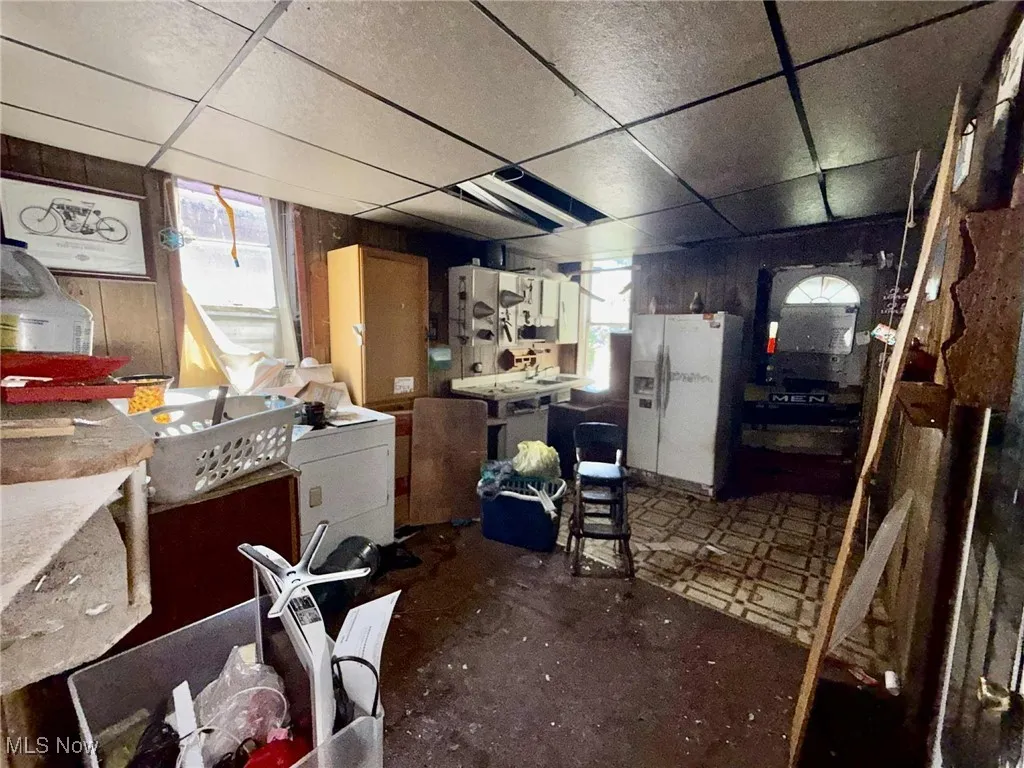 Kitchen featuring a drop ceiling, wooden walls, freestanding refrigerator, white cabinetry, and washer / dryer