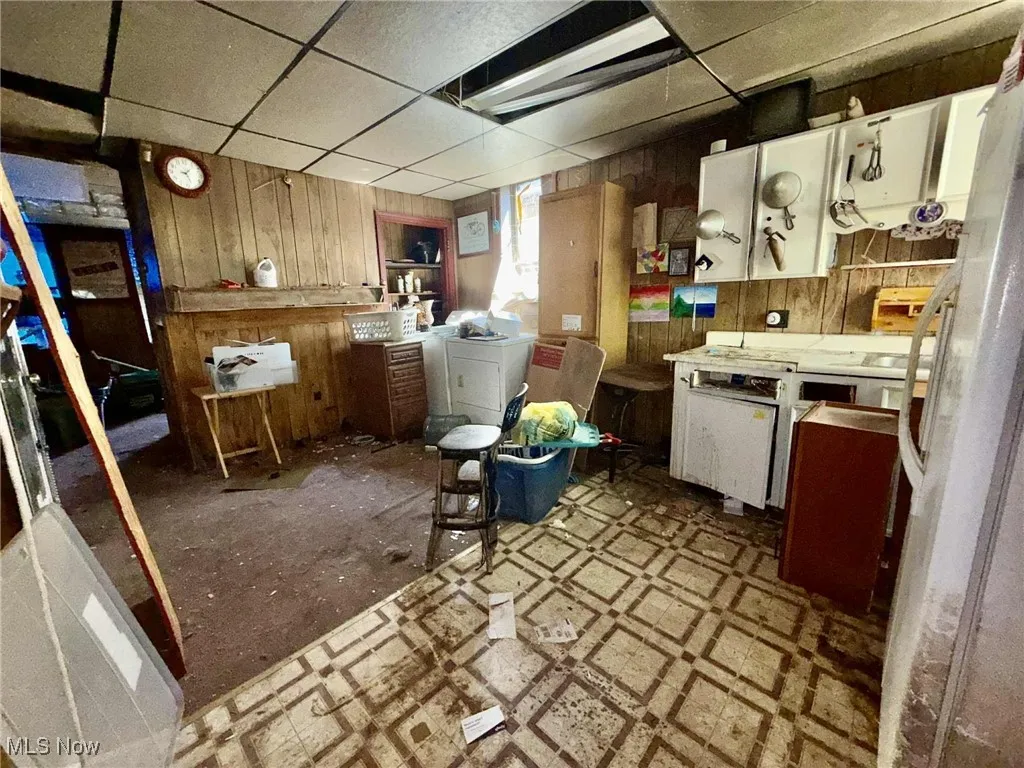 Kitchen featuring a drop ceiling, wooden walls, freestanding refrigerator, white cabinetry, and washer / dryer