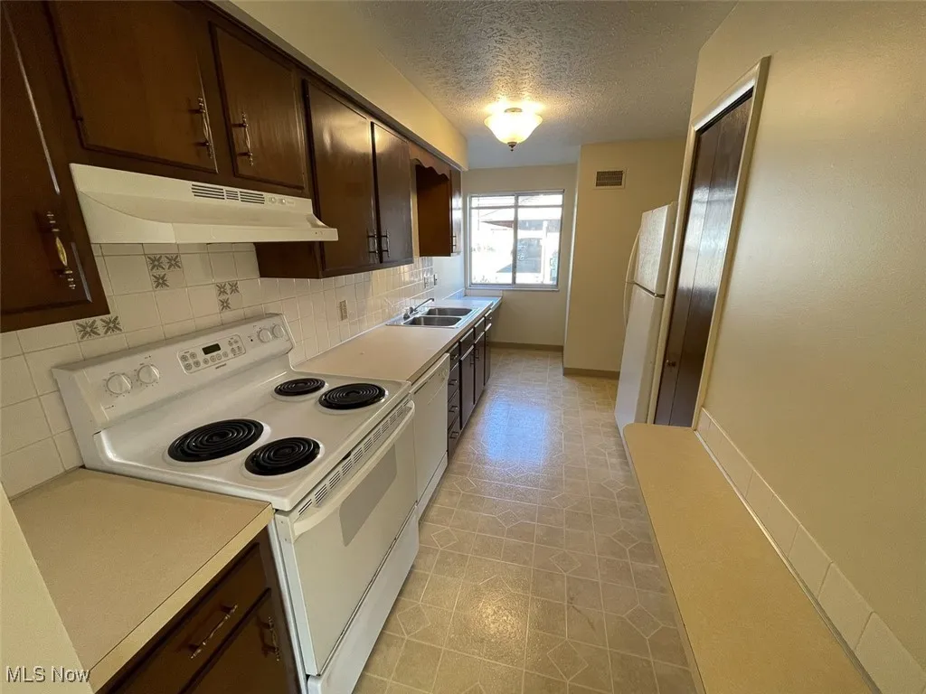 Kitchen with dark brown cabinetry, white appliances, light countertops, backsplash, and under cabinet range hood