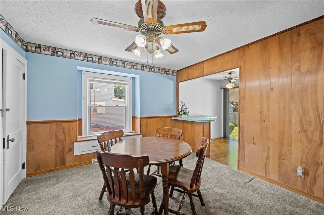 Dining space with wooden walls, a textured ceiling, a wainscoted wall, carpet floors, and a ceiling fan