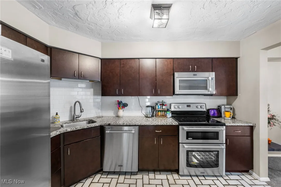 Kitchen featuring dark brown cabinets, stainless steel appliances, tasteful backsplash, light stone countertops, and a textured ceiling