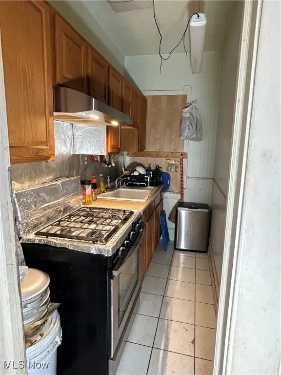 Kitchen featuring decorative backsplash, stainless steel range, brown cabinetry, extractor fan, and light tile patterned flooring