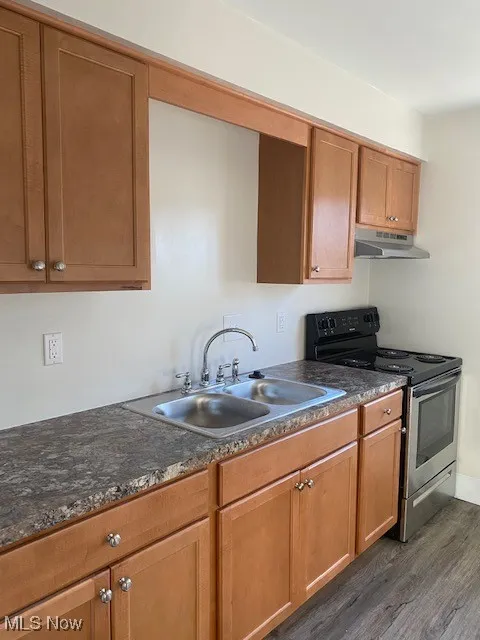 Kitchen featuring stainless steel electric range oven, brown cabinetry, dark wood-type flooring, under cabinet range hood, and dark stone counters