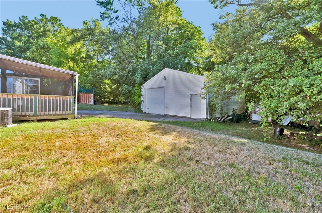 View of grassy yard featuring an outdoor structure, driveway, a detached garage, view of wooded area, and a sunroom