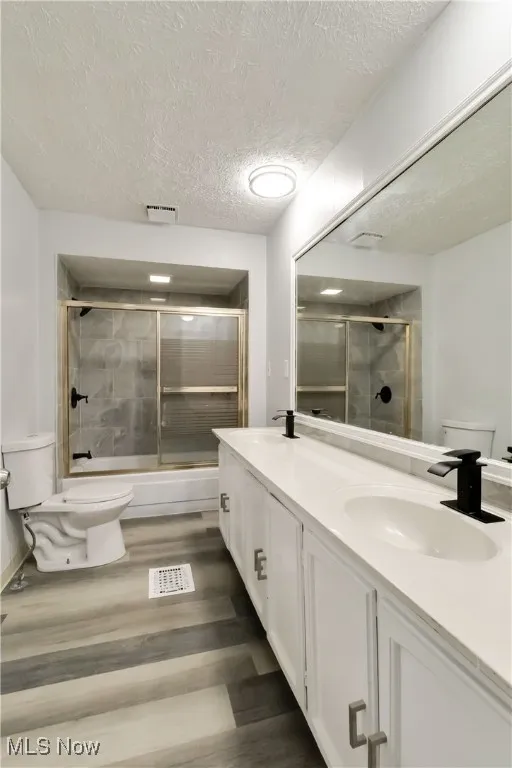 Full bath with combined bath / shower with glass door, double vanity, dark wood-type flooring, and a textured ceiling