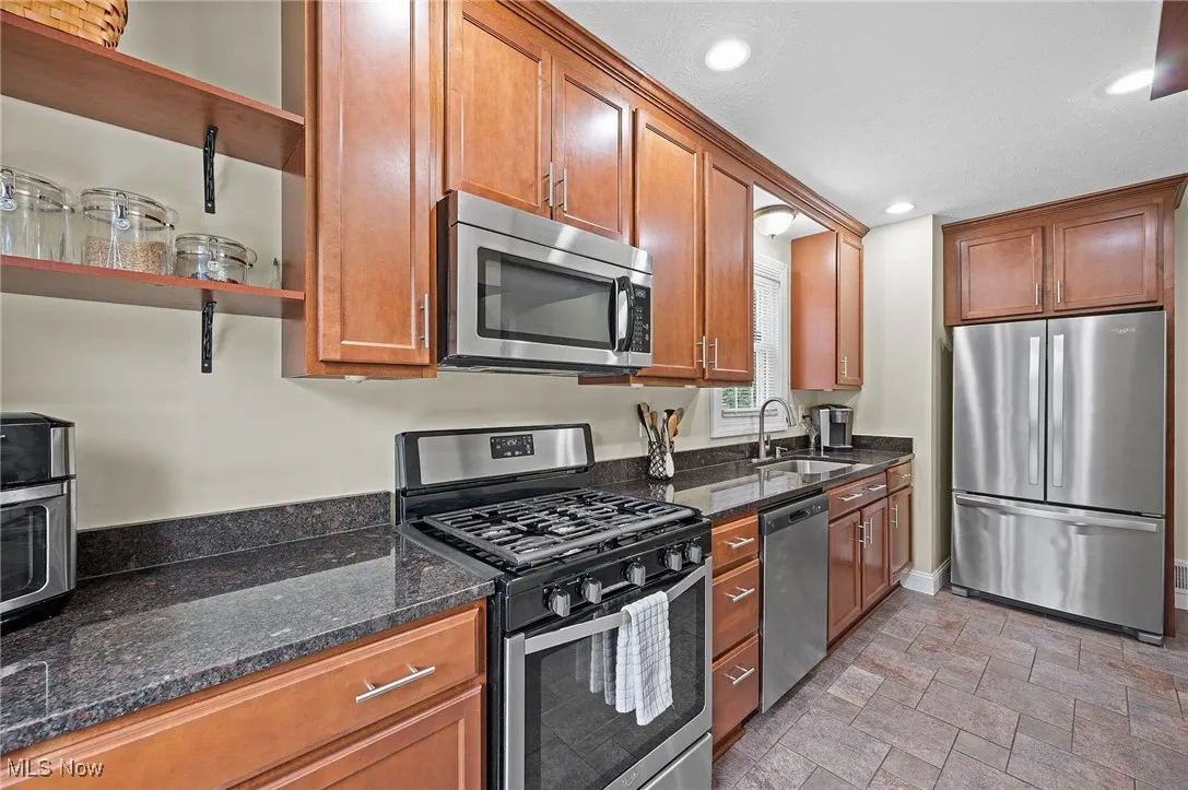 Kitchen featuring appliances with stainless steel finishes, open shelves, dark stone countertops, brown cabinetry, and light stone finish floors
