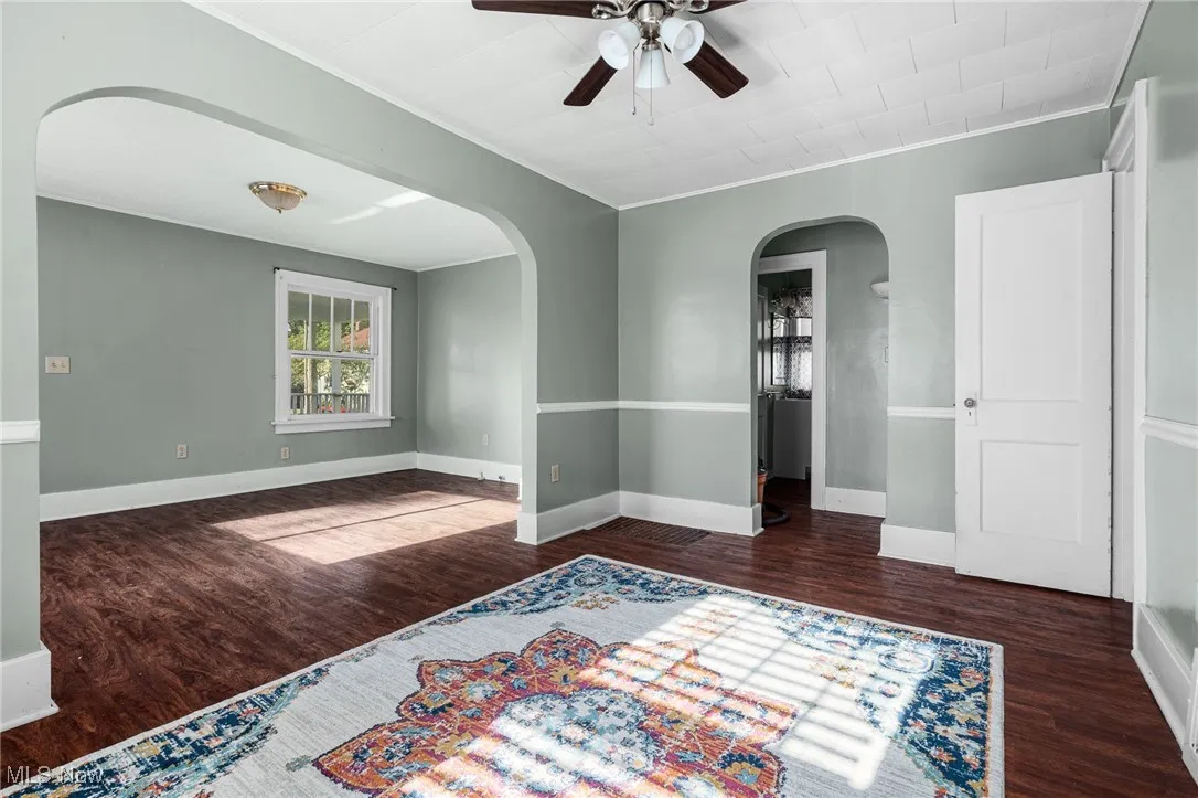 Empty room with arched walkways, dark wood finished floors, a ceiling fan, and ornamental molding