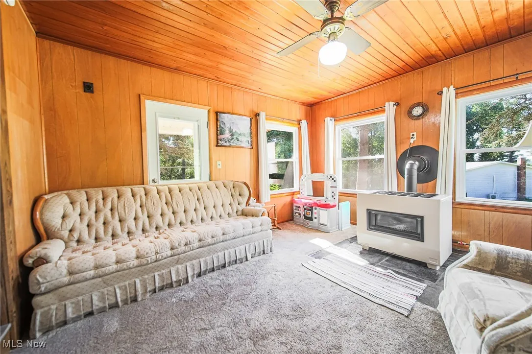 Carpeted living room with a wood stove, healthy amount of natural light, wooden ceiling, wooden walls, and ceiling fan
