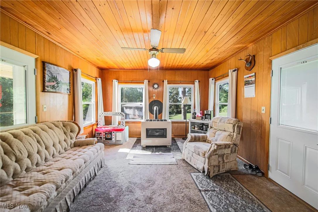 Living area featuring wooden walls, a wood stove, wooden ceiling, a ceiling fan, and carpet