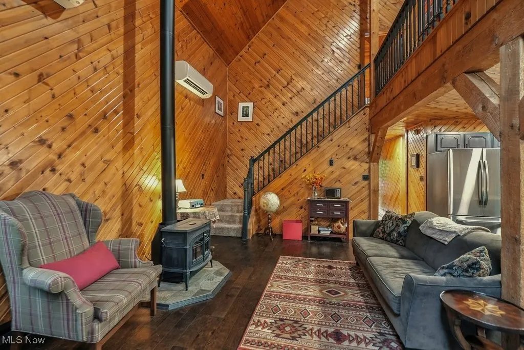 Living area with wooden walls, a wood stove, stairs, high vaulted ceiling, and dark wood-type flooring