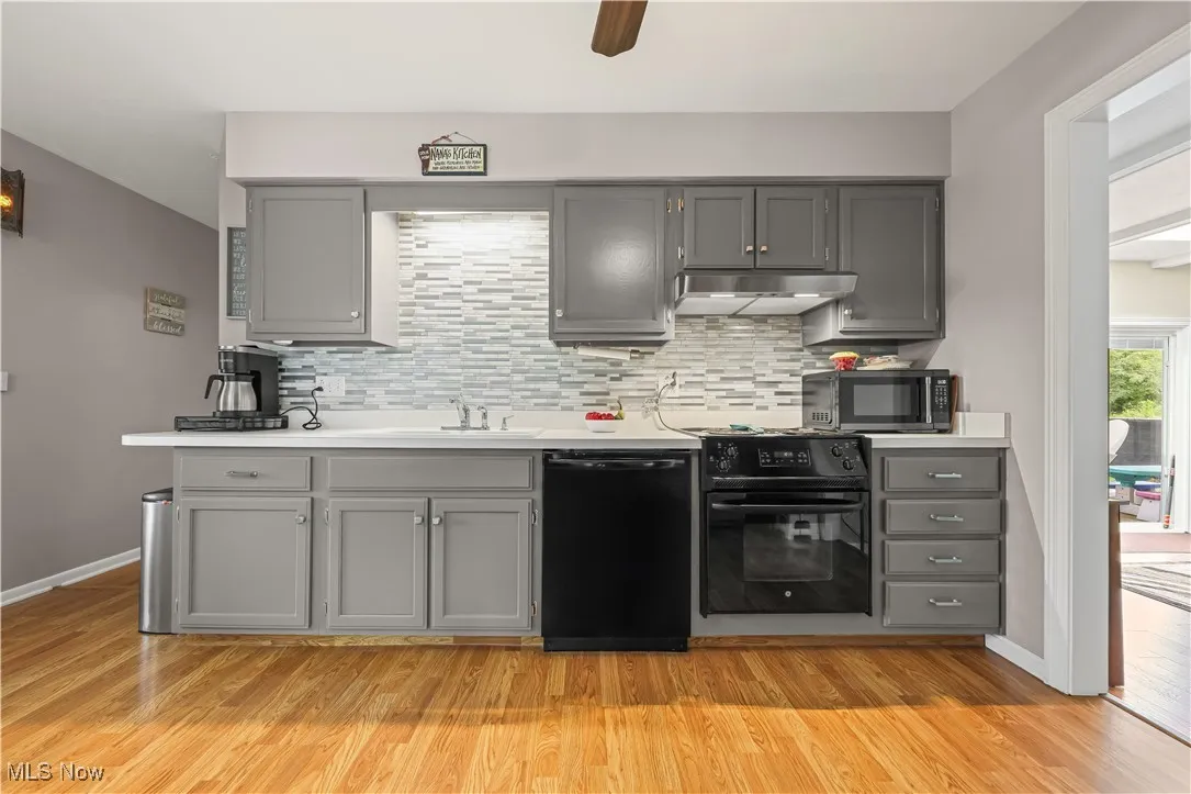 Kitchen with gray cabinets, decorative backsplash, black appliances, light countertops, and ceiling fan