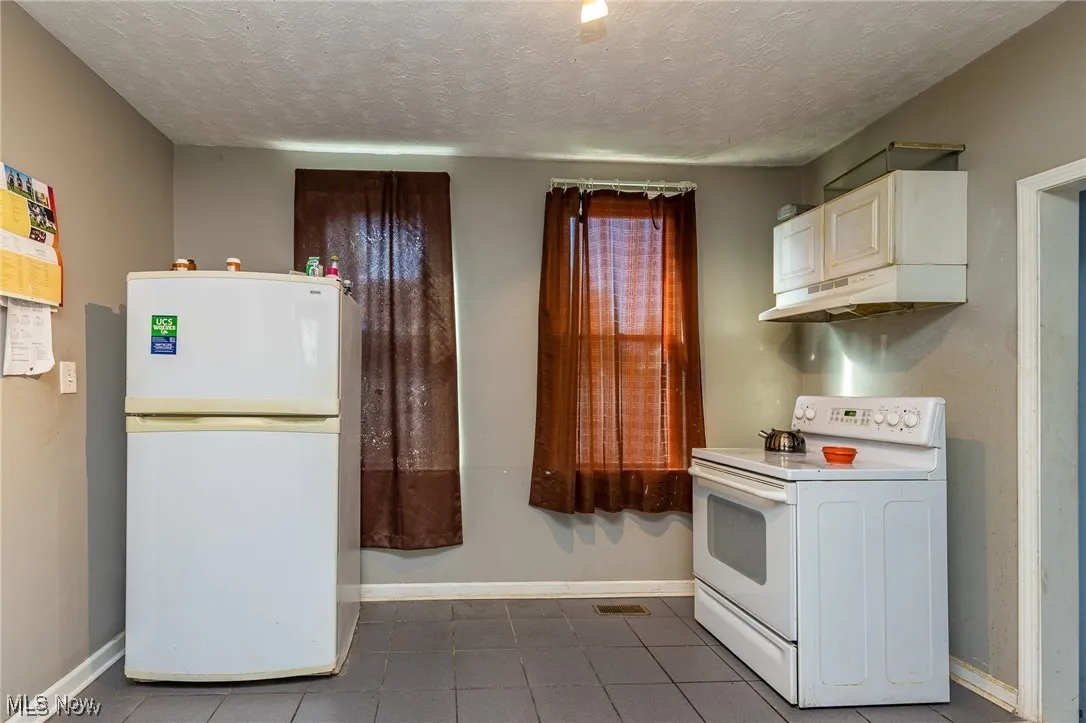 Kitchen with white appliances, under cabinet range hood, a textured ceiling, dark tile patterned floors, and white cabinetry