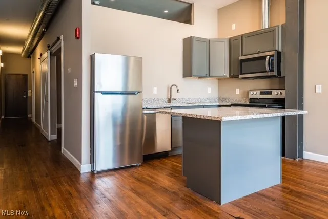 Kitchen with stainless steel appliances, gray cabinets, light stone countertops, a kitchen island, and dark wood-style flooring