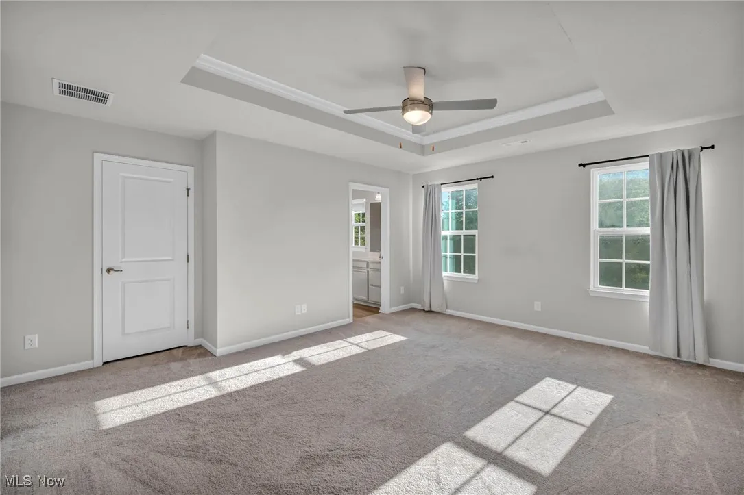 Unfurnished bedroom featuring a tray ceiling, light colored carpet, a ceiling fan, ensuite bath, and ornamental molding