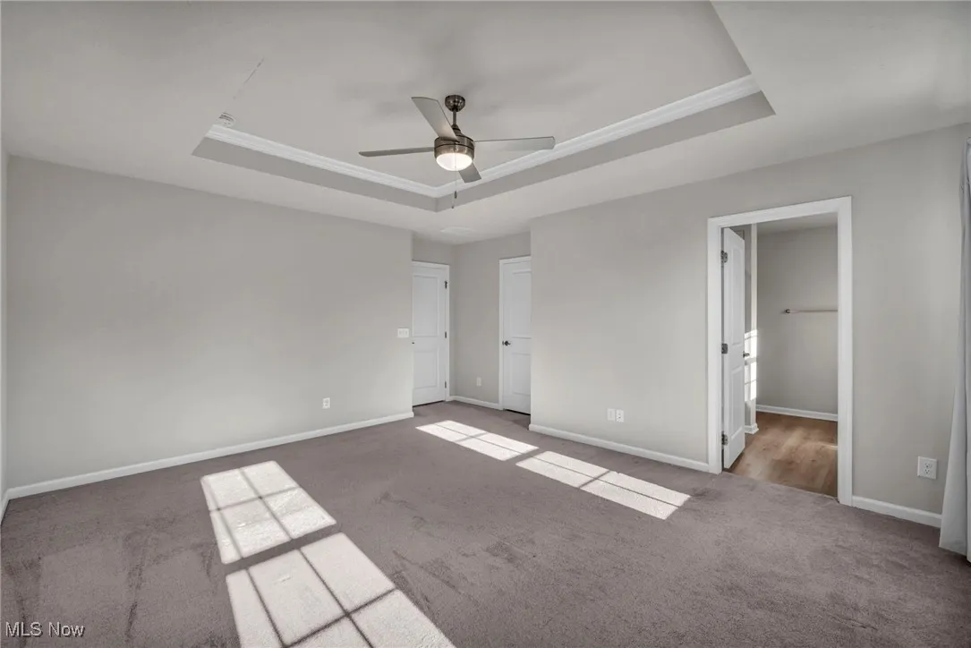 Unfurnished bedroom featuring a tray ceiling, carpet flooring, ceiling fan, ensuite bath, and ornamental molding