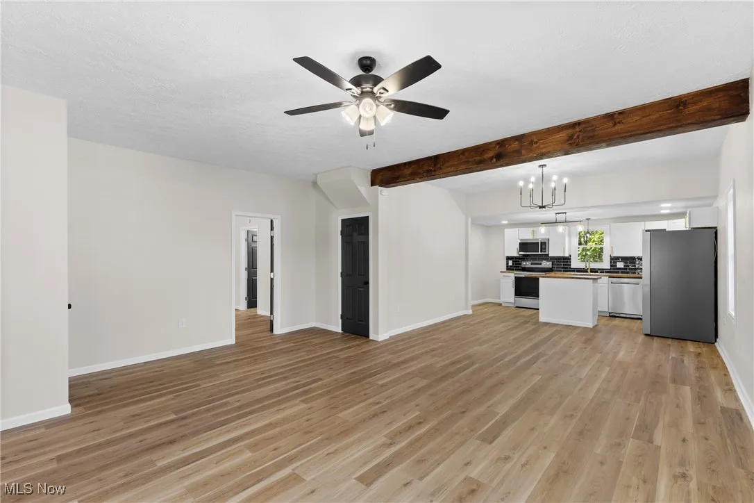 Unfurnished living room featuring beamed ceiling, light wood-style floors, a chandelier, and a ceiling fan
