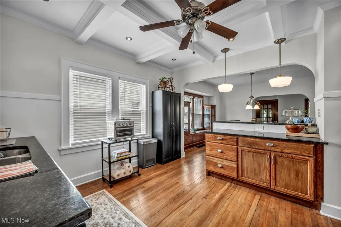 Kitchen with beamed ceiling, arched walkways, brown cabinetry, light wood-style flooring, and hanging light fixtures