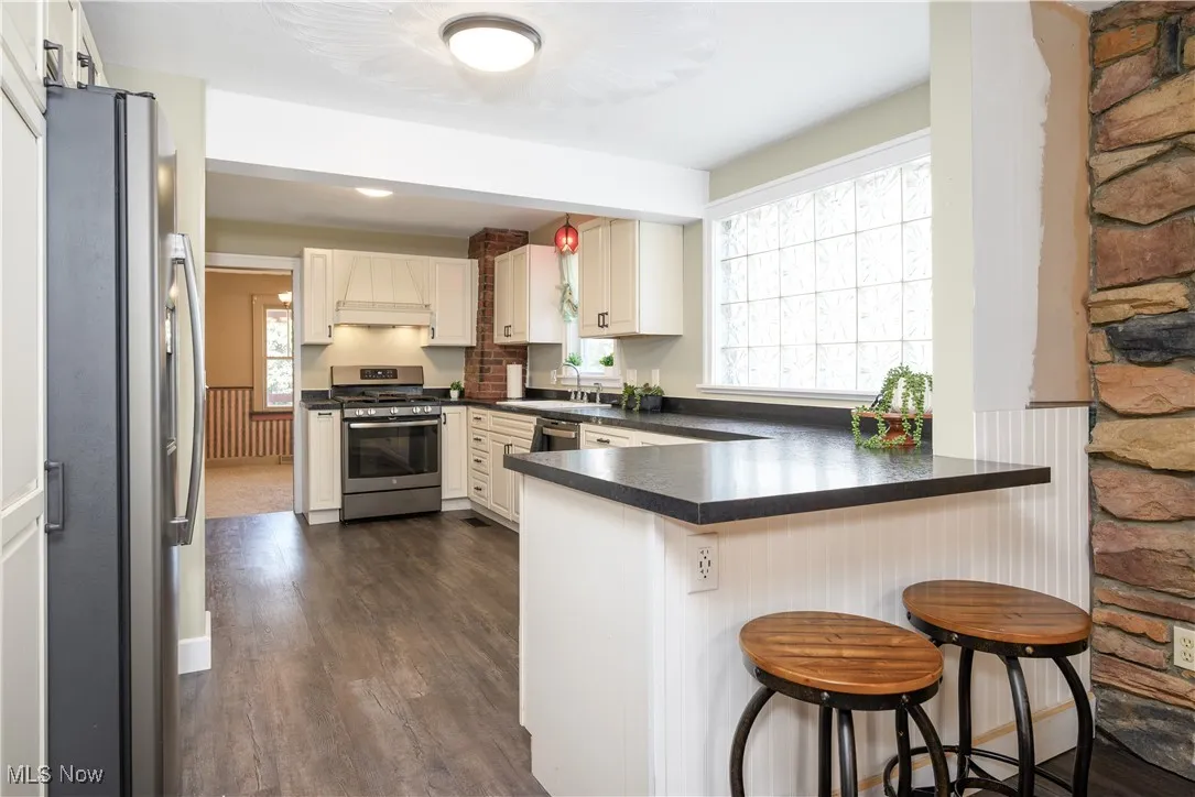 Kitchen with a peninsula, stainless steel appliances, dark countertops, premium range hood, and a breakfast bar
