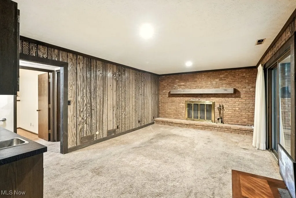Unfurnished living room with light colored carpet, wooden walls, a brick fireplace, crown molding, and a textured ceiling