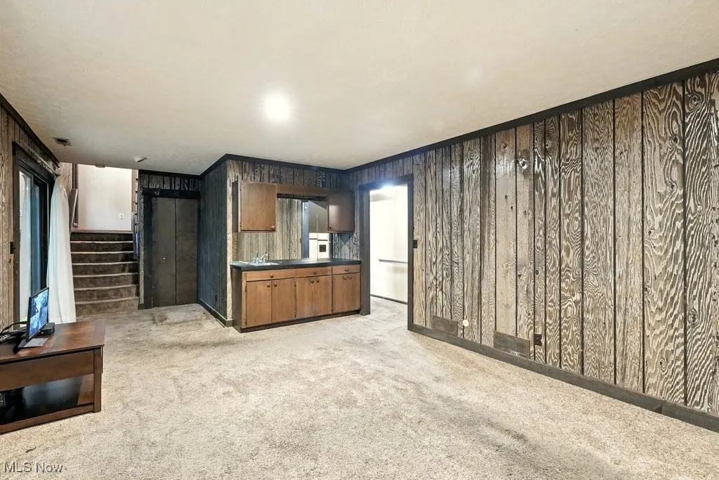 Unfurnished living room featuring light colored carpet, wooden walls, and stairs