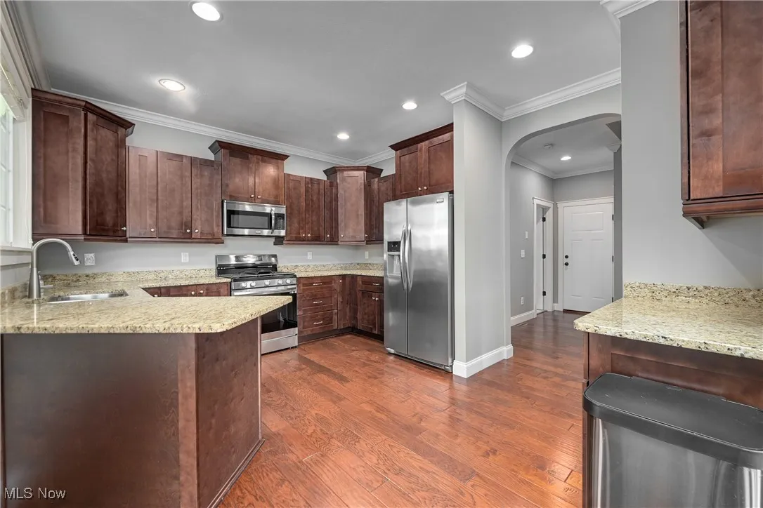 Kitchen with arched walkways, appliances with stainless steel finishes, light stone counters, dark brown cabinets, and ornamental molding