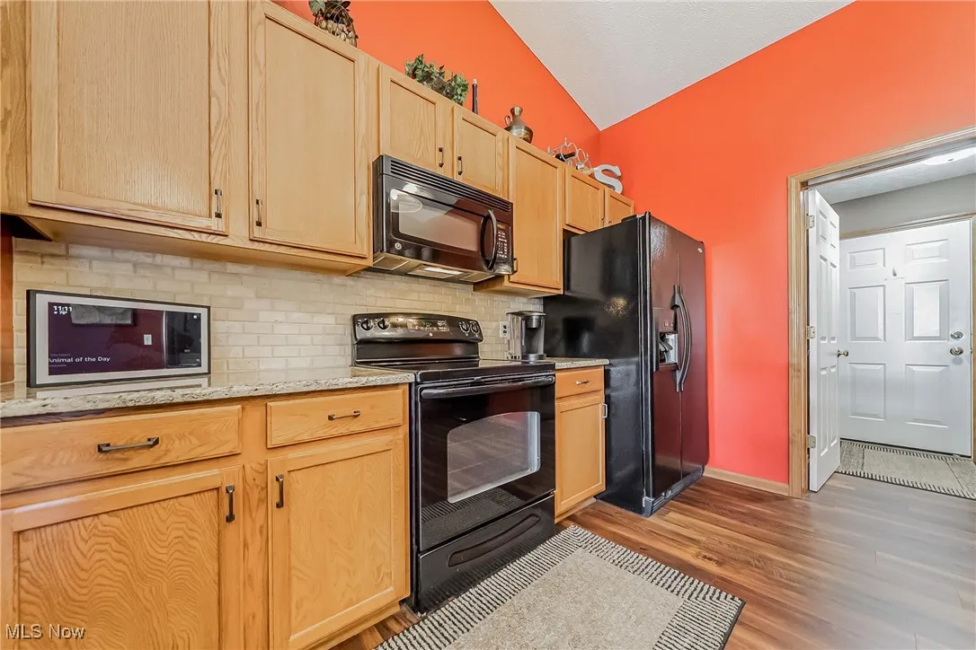 Kitchen with black appliances, decorative backsplash, light stone counters, dark wood-style flooring, and light brown cabinetry