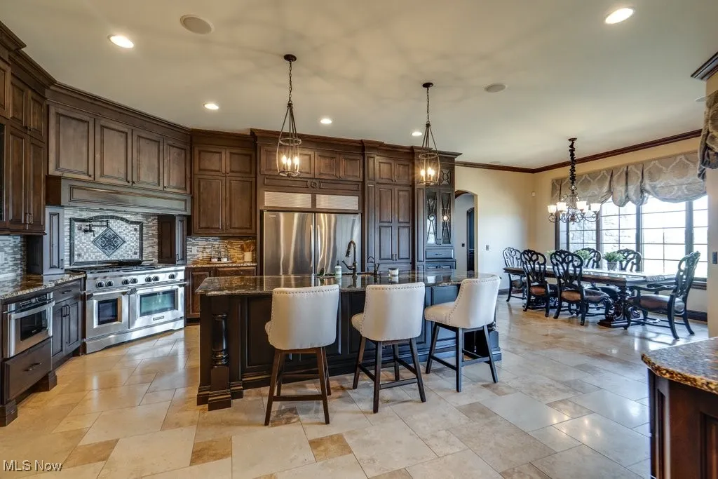 Kitchen featuring arched walkways, dark brown cabinets, dark stone countertops, hanging light fixtures, and crown molding