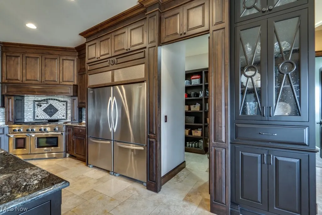 Kitchen with appliances with stainless steel finishes, dark stone countertops, glass insert cabinets, dark brown cabinets, and recessed lighting