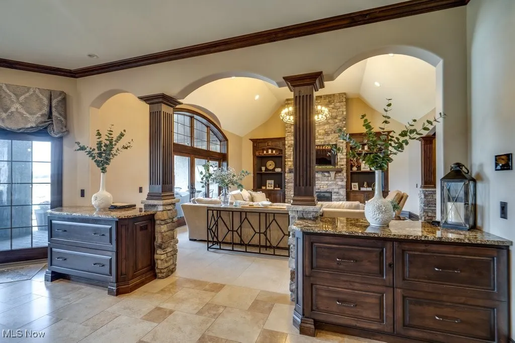 Kitchen featuring dark stone countertops, arched walkways, dark brown cabinets, decorative columns, and high vaulted ceiling