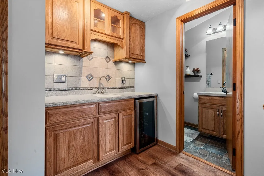 Indoor wet bar with backsplash, beverage cooler, glass insert cabinets, dark wood-style flooring, and brown cabinetry