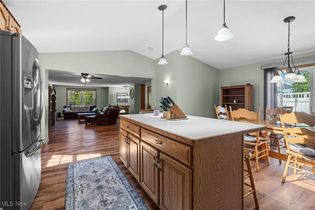 Kitchen with brown cabinets, stainless steel fridge with ice dispenser, hanging light fixtures, dark wood finished floors, and vaulted ceiling