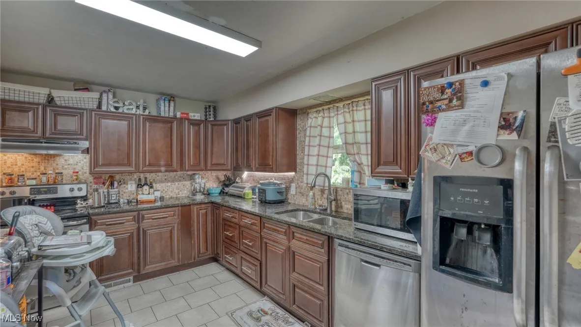 Kitchen with appliances with stainless steel finishes, dark stone countertops, light tile patterned floors, tasteful backsplash, and under cabinet range hood