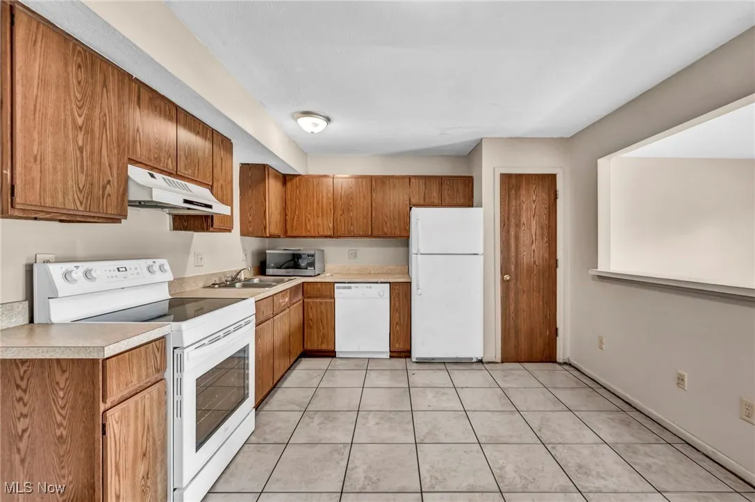 Kitchen featuring white appliances, brown cabinets, light countertops, under cabinet range hood, and light tile patterned floors