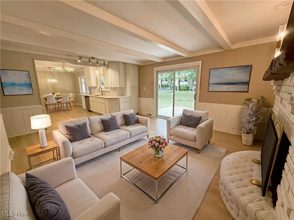 Living area featuring a wainscoted wall, light wood-style floors, beamed ceiling, a stone fireplace, and a chandelier