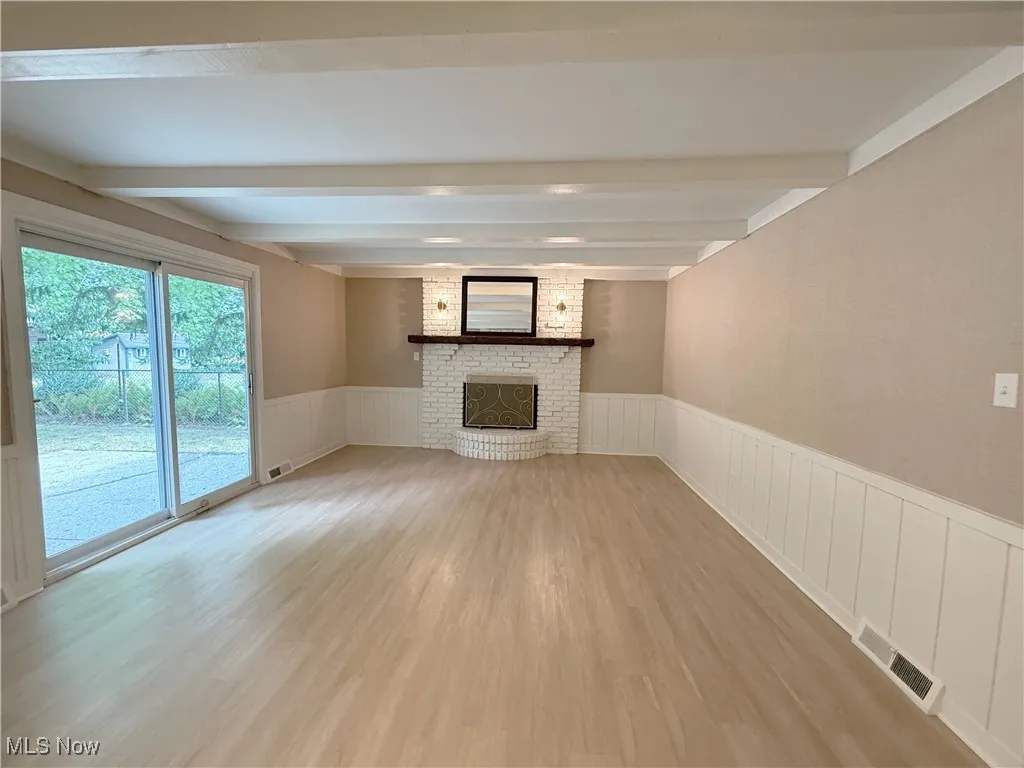 Unfurnished living room with a wainscoted wall, light wood finished floors, a fireplace, beam ceiling, and a decorative wall