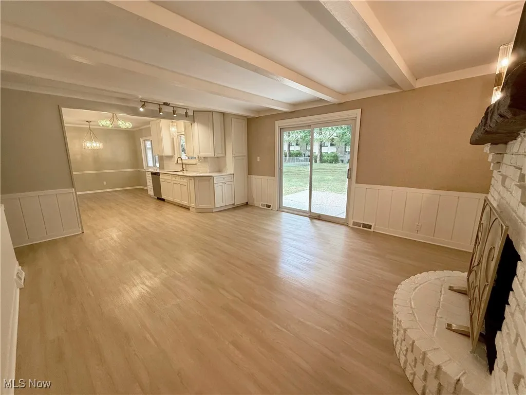 Unfurnished living room featuring wainscoting, a fireplace with raised hearth, light wood finished floors, a chandelier, and beam ceiling