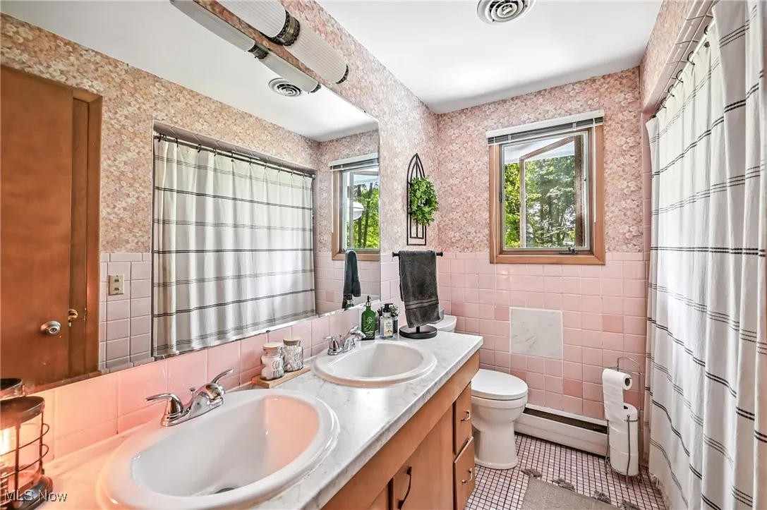 Bathroom featuring wallpapered walls, tile walls, double vanity, baseboard heating, and light tile patterned flooring