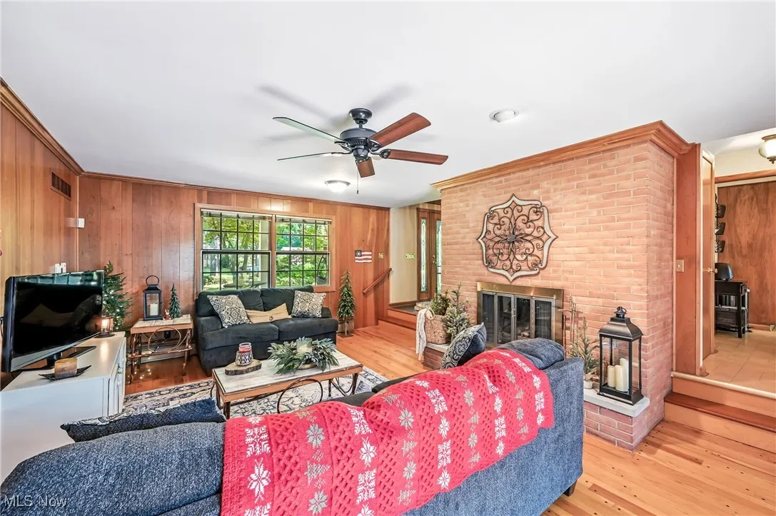 Living area featuring wooden walls, crown molding, hardwood floors, a brick fireplace, and ceiling fan