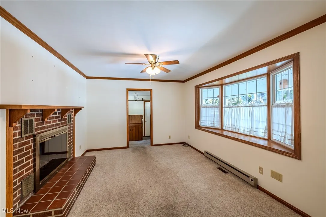 Unfurnished living room with ornamental molding, light colored carpet, a baseboard radiator, a brick fireplace, and a ceiling fan