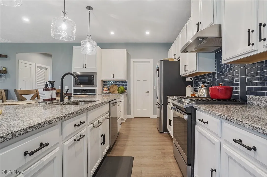 Kitchen featuring white cabinetry, stainless steel appliances, light stone counters, under cabinet range hood, and hanging light fixtures
