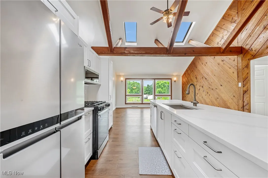 Kitchen featuring stainless steel appliances, white cabinets, wood walls, beamed ceiling, and a skylight