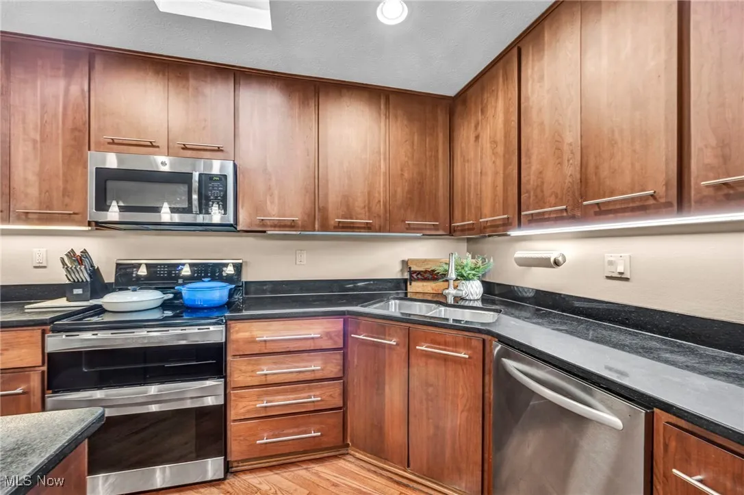 Kitchen featuring stainless steel appliances, light wood-style floors, brown cabinetry, dark stone countertops, and a textured ceiling