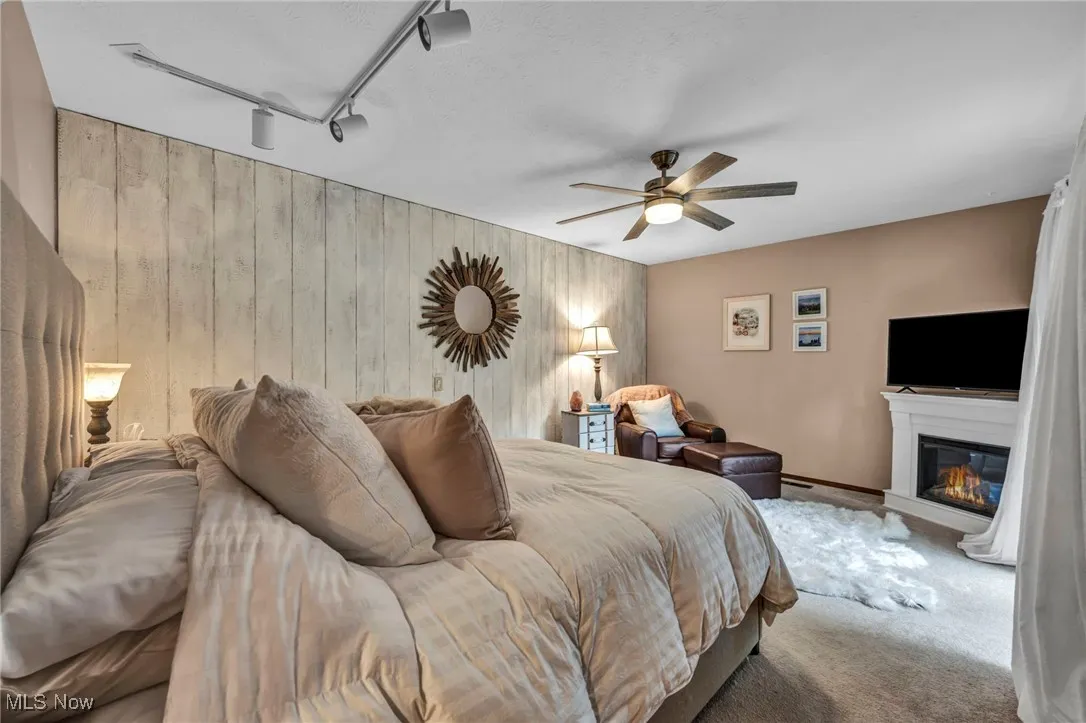 Carpeted bedroom featuring rail lighting, wooden walls, a glass covered fireplace, ceiling fan, and a textured ceiling