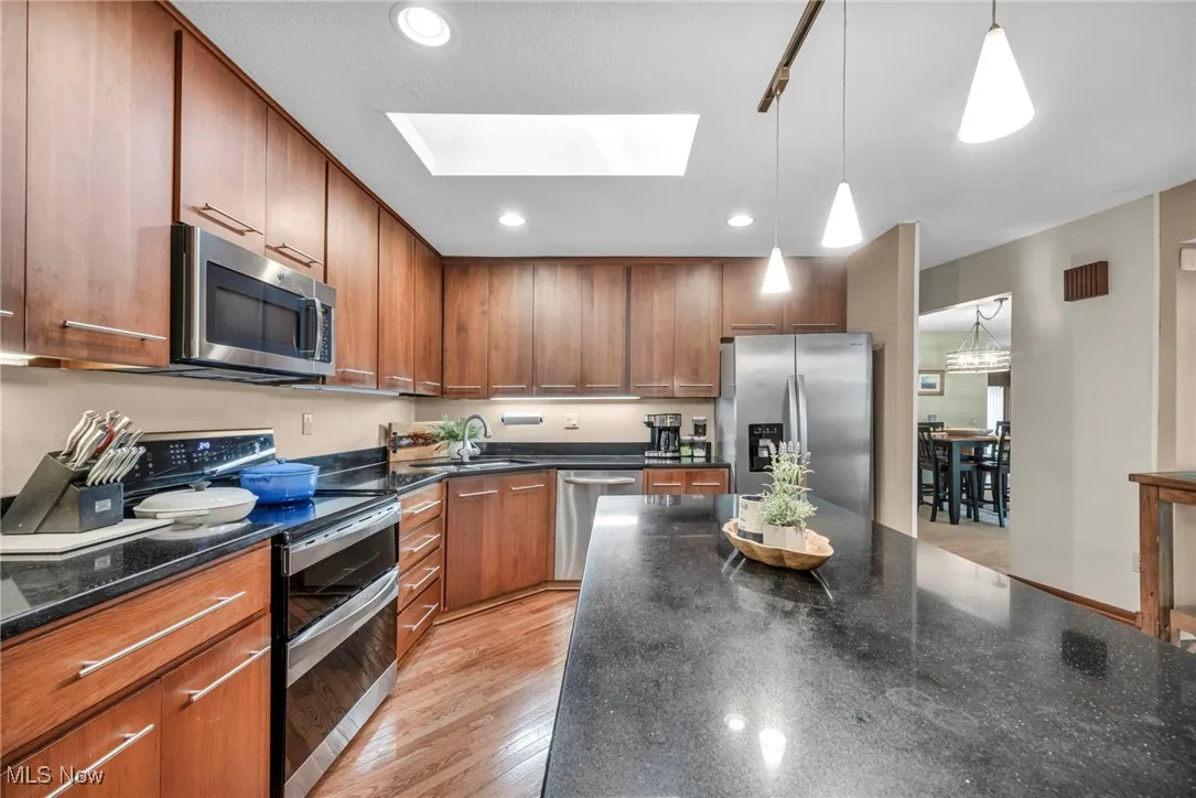 Kitchen with appliances with stainless steel finishes, dark stone counters, light wood finished floors, decorative light fixtures, and brown cabinetry
