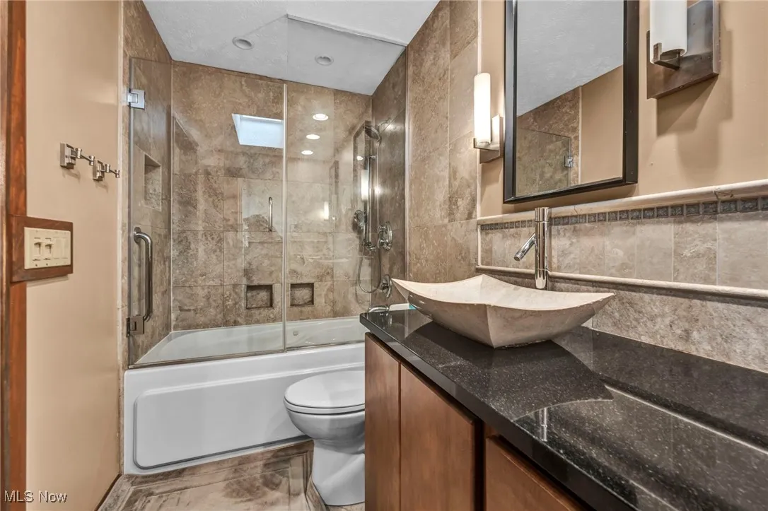 Full bath with combined bath / shower with glass door, double vanity, tile walls, and a skylight