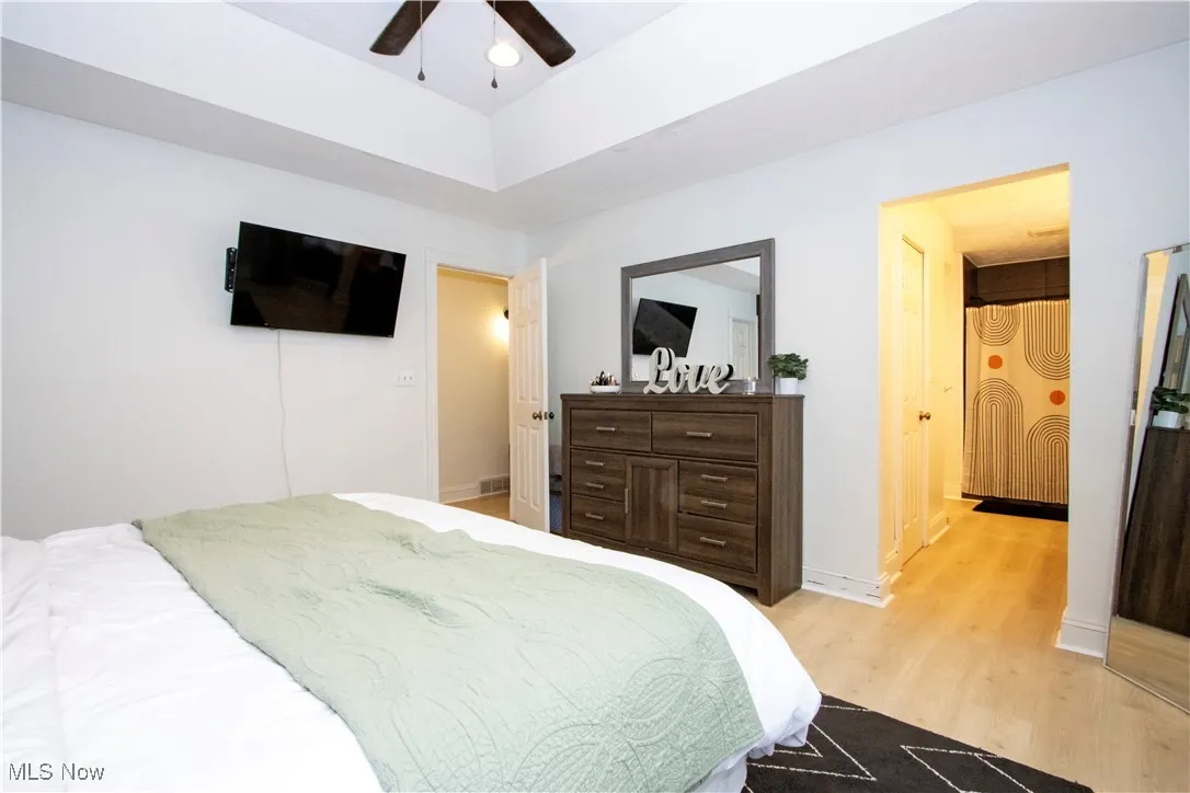 Bedroom featuring light wood-style flooring, ensuite bath, a ceiling fan, and a raised ceiling