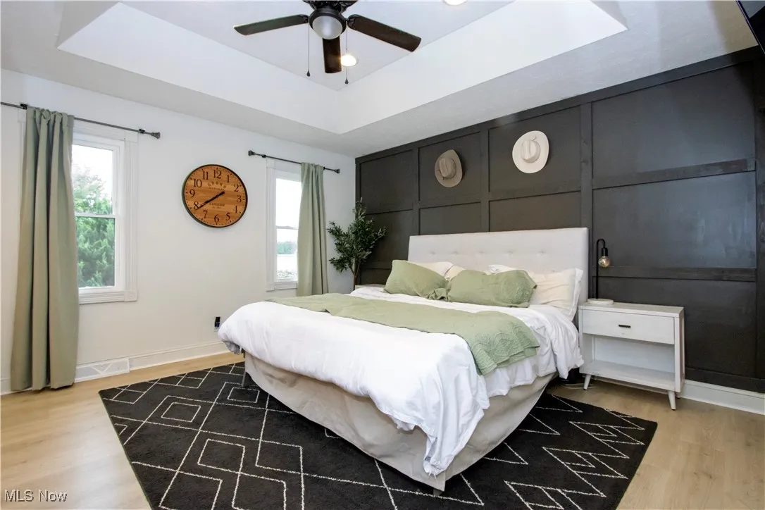 Bedroom featuring a tray ceiling, multiple windows, light wood-type flooring, and a ceiling fan