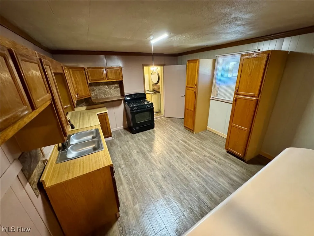 Kitchen with crown molding, light wood-type flooring, black range with gas cooktop, brown cabinetry, and light countertops