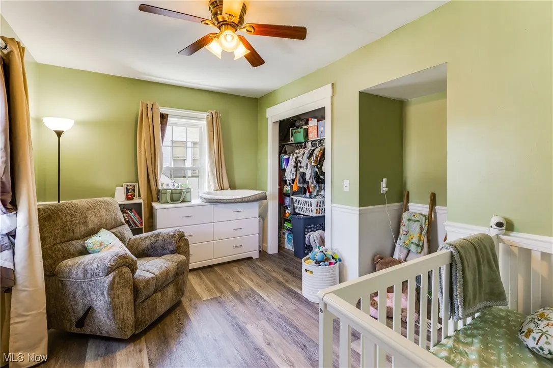 Bedroom with wood finished floors, a nursery area, a closet, ceiling fan, and wainscoting