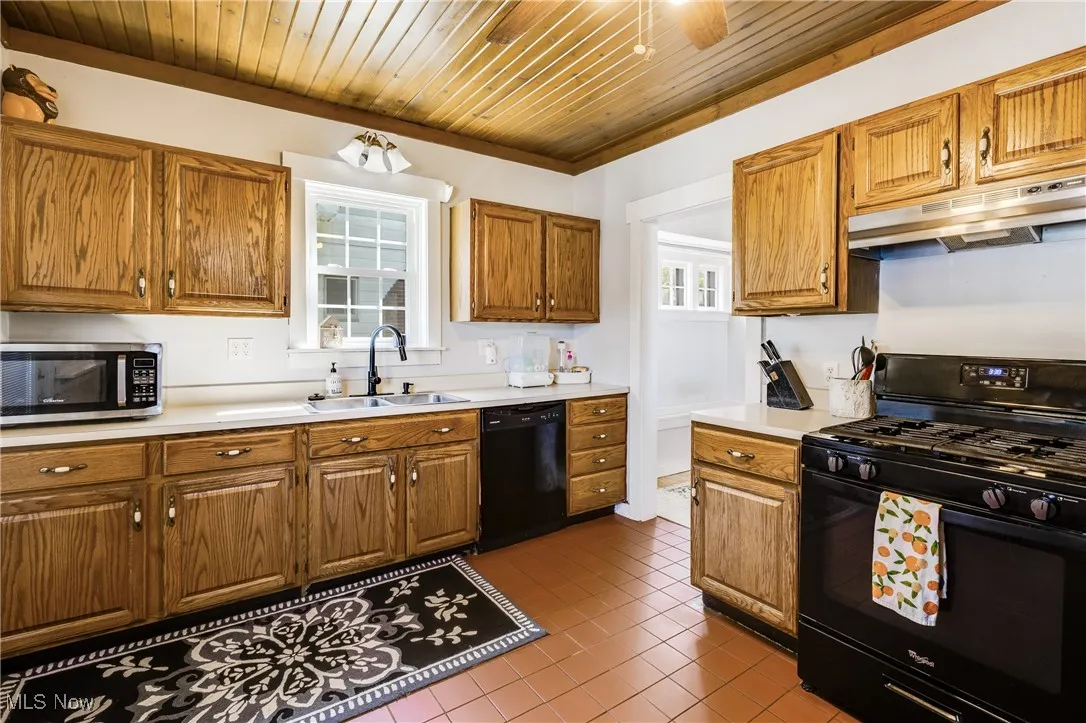 Kitchen featuring black appliances, light countertops, brown cabinetry, under cabinet range hood, and dark tile patterned floors